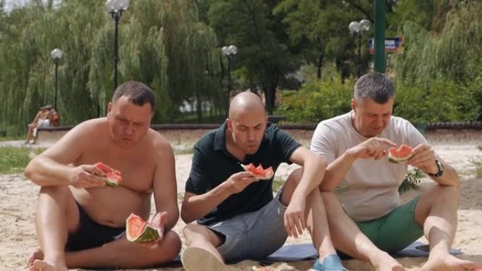 一群朋友在夏天吃西瓜的野餐时间。三个男性朋友非常有魅力和可爱坐在草地上野餐。慢动作