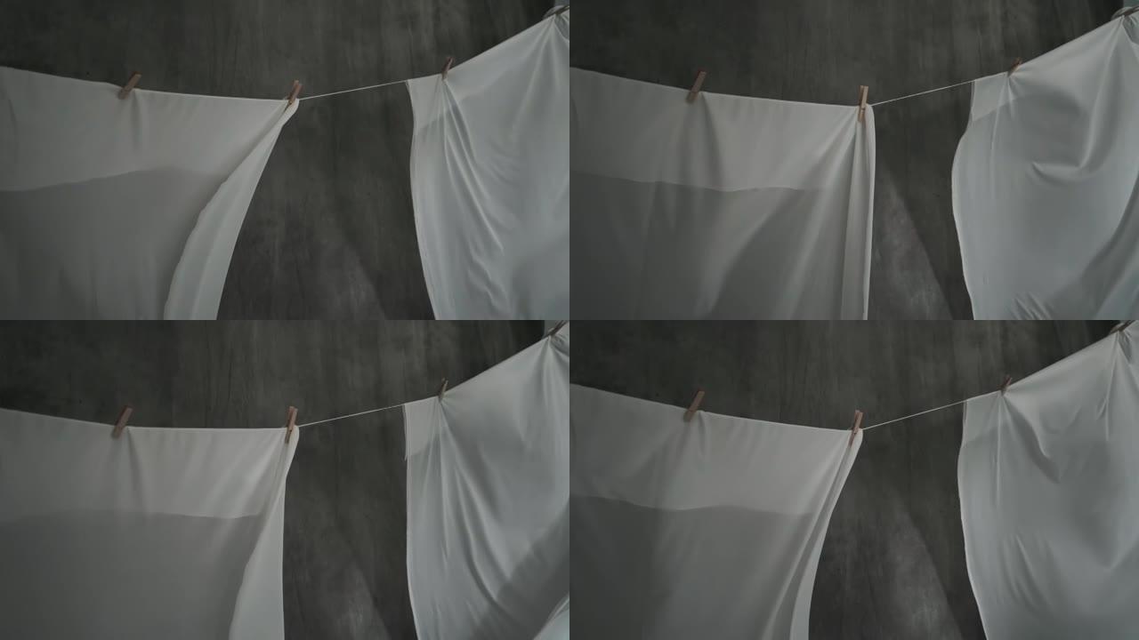 白色织物床单在绳子上的衣夹上干燥。波浪织物在风中飘动。抽象灰色背景。