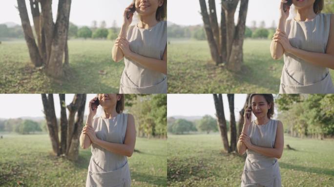 年轻美丽的晒黑皮肤女孩拿着智能手机走在公园内的大树下，漫步在新鲜的绿色广阔的草地上，夏天的阳光普照，