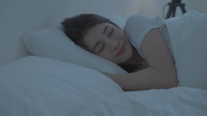 穿着睡衣的亚洲美丽舒适女孩在黑暗的夜晚睡在卧室里。迷人的年轻女子躺在床上，用舒适的枕头和毯子感到幸福