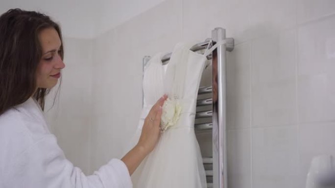 挂在浴室的婚纱侧视图，微笑着快乐自信的年轻女子进入触摸白色优雅奢华的衣服。婚礼早晨快乐的新娘欣赏礼服