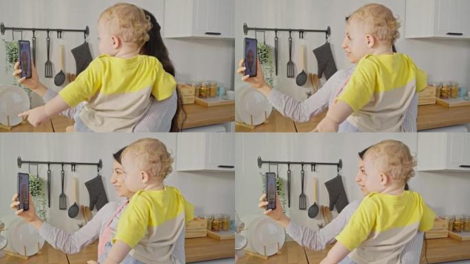 白人母亲抱着小婴儿，与家人在线视频通话。迷人的漂亮妈妈用手机和丈夫聊天，同时带着年幼的蹒跚学步的孩子