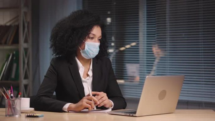 戴着医用口罩的非洲裔美国妇女的肖像在笔记本电脑上的视频会议上讲话。女商人在工作场所摆姿势，坐在办公室