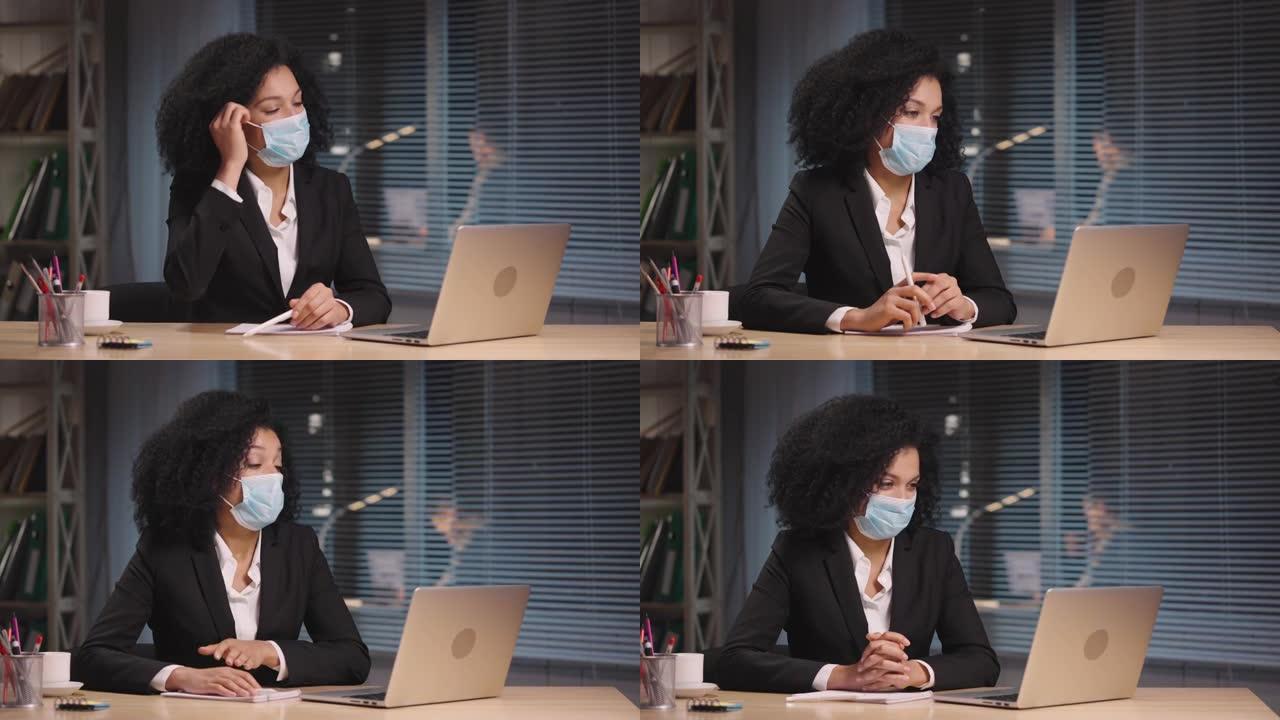戴着医用口罩的非洲裔美国妇女的肖像在笔记本电脑上的视频会议上讲话。女商人在工作场所摆姿势，坐在办公室