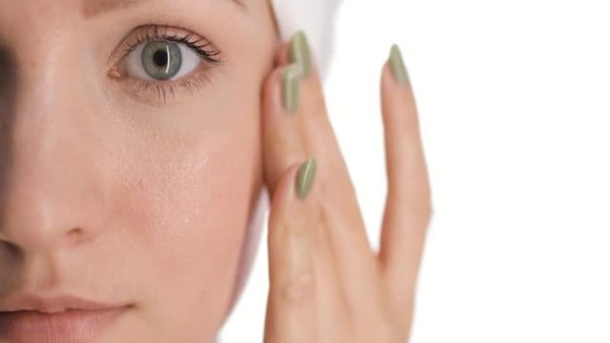 欧洲年轻女子脸上的一部分特写，头上抹着干发毛巾，用手轻敲和微笑在脸颊上涂抹护肤霜