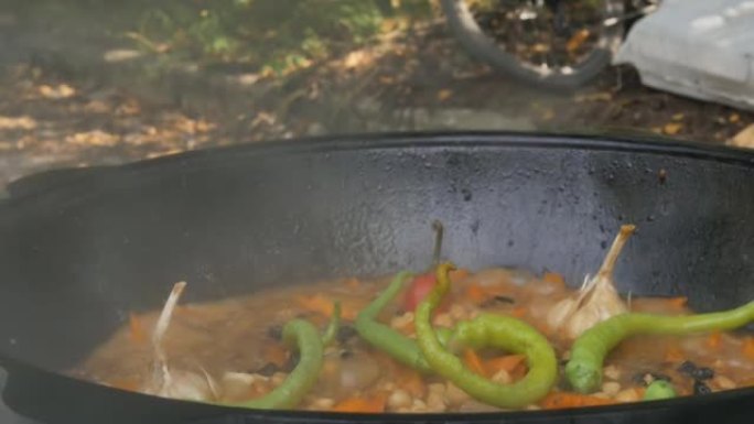 厨师的手用烹饪乌兹别克抓饭抬起大锅的盖子。令人垂涎的蔬菜在露天大锅中煮沸并飙升。大蒜的头以及红色和绿