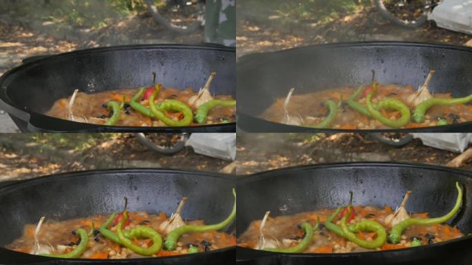 厨师的手用烹饪乌兹别克抓饭抬起大锅的盖子。令人垂涎的蔬菜在露天大锅中煮沸并飙升。大蒜的头以及红色和绿