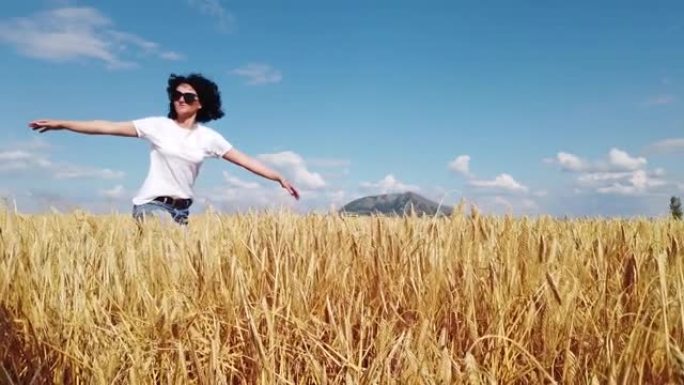 夏日晴天，黑色卷发的快乐迷人女人的慢动作走在金色的麦田里