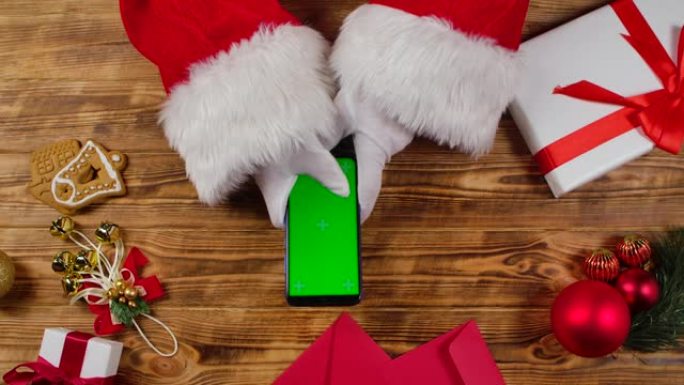 顶视图圣诞老人手使用智能手机与绿色屏幕色度键由木制新年装饰桌。圣诞老人在屏幕上点击观看内容，进行在线