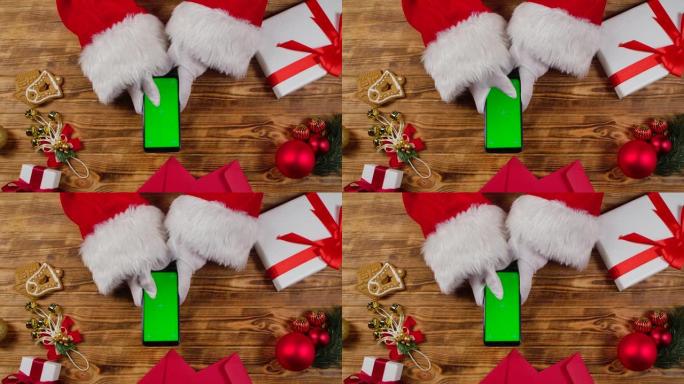 顶视图圣诞老人手使用智能手机与绿色屏幕色度键由木制新年装饰桌。圣诞老人在屏幕上点击观看内容，进行在线
