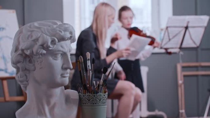 年轻的金发女老师给班上的一个小女孩上小提琴课 -- 希腊男人的半身像和艺术画笔放在前景上