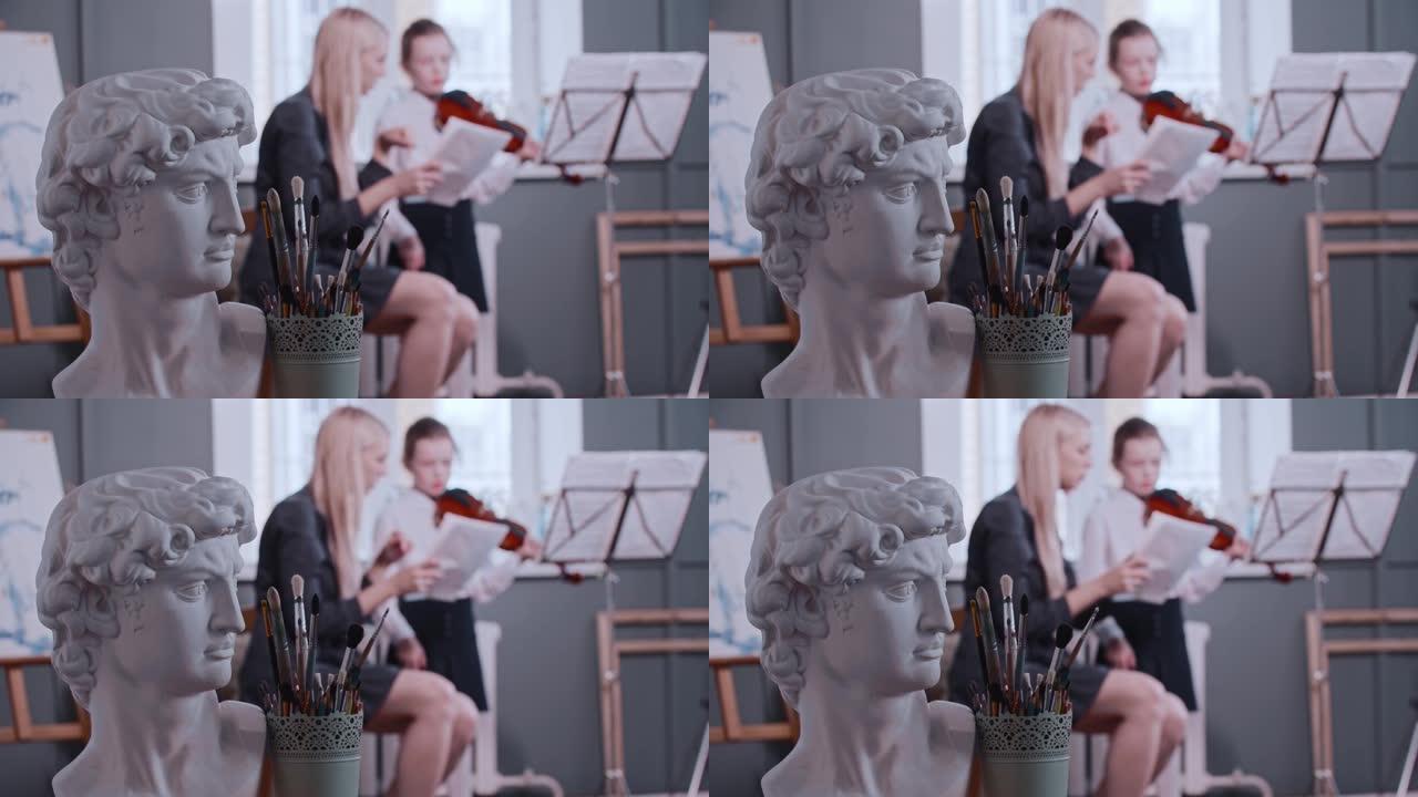 年轻的金发女老师给班上的一个小女孩上小提琴课 -- 希腊男人的半身像和艺术画笔放在前景上