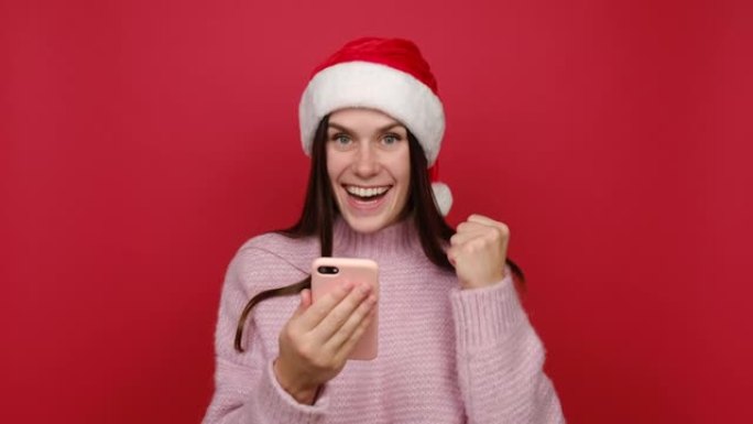 年轻的圣诞老人高加索女人拿着的肖像用手机打字说哇是的刚刚发现了大胜利新闻，穿着粉色毛衣和圣诞帽，孤立