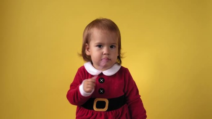 可爱的快乐快乐胖乎乎的学步女婴，穿着圣诞老人套装，在黄色背景下看着相机。庆祝生日的孩子玩耍场景。孩子