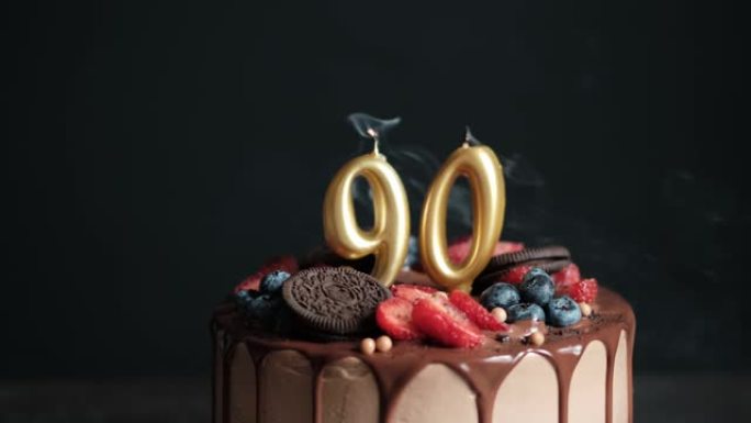 在黑色背景的巧克力生日蛋糕上吹灭蜡烛，上面有90号金色蜡烛。90周年生日蛋糕。慢动作