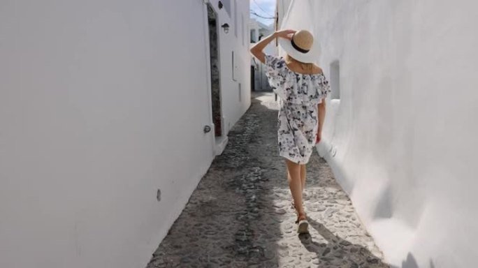 戴着草帽和优雅连衣裙的女人走在圣托里尼岛的街道上。希腊