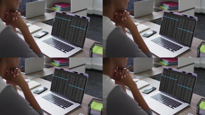 混血女人坐在办公桌前看着笔记本电脑屏幕上的编码数据处理