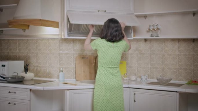 穿着绿色连衣裙的女孩把东西放在厨柜里，整理盘子，后视图