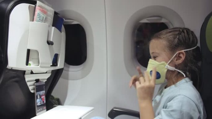 戴着防护口罩的白人小女孩。带呼吸器的飞机上的儿童婴儿游客在智能手机上玩耍。新型冠状病毒肺炎。