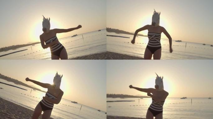 穿着独角兽面具的有趣女人黎明时在海滩上跳舞。泳装和独角兽头女人的快乐舞蹈。假期快乐。摄像机运动。