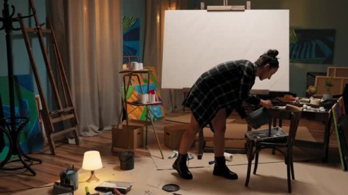 一个画家的工作室，在中间矗立着一个画架，画架上有大画布，旁边是美丽的女人弯下腰去拿一罐灰色油漆，倒入