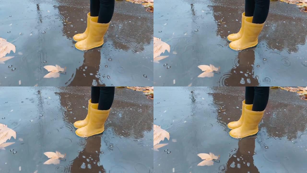 穿着黄色胶鞋的孩子站在水坑里，靠近地面上的秋叶