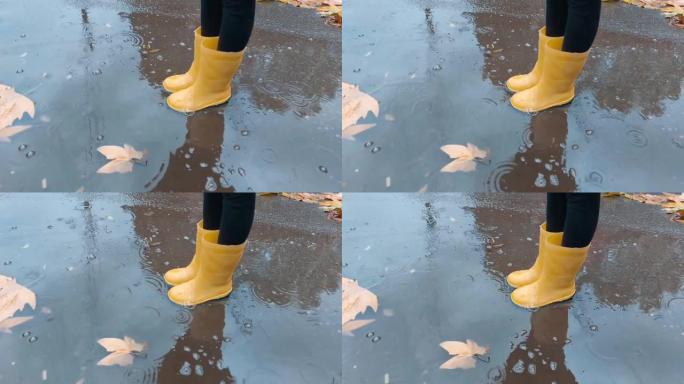 穿着黄色胶鞋的孩子站在水坑里，靠近地面上的秋叶