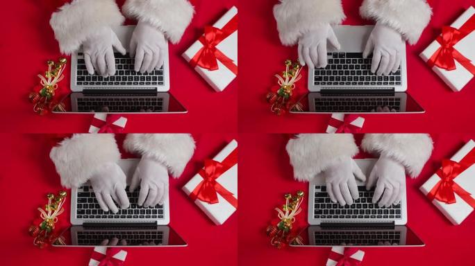 顶视图圣诞老人戴着白手套在红色新年装饰桌的键盘上打字。圣诞老人与笔记本电脑一起工作，浏览邮件并回答给