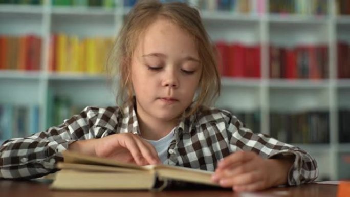 严肃的小学生小学生的特写镜头前视图翻过书页，坐在桌子旁看书，背景模糊。
