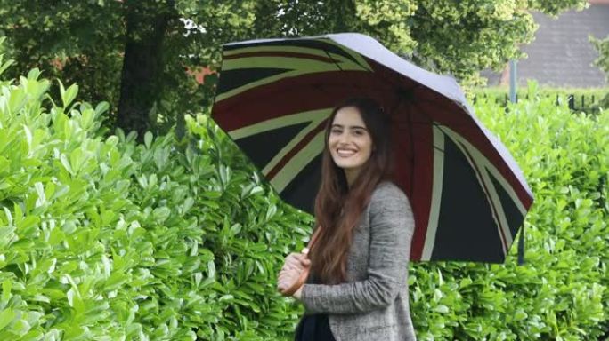 一名年轻女子打开英国国旗伞，在肩膀上旋转，看着相机，微笑着