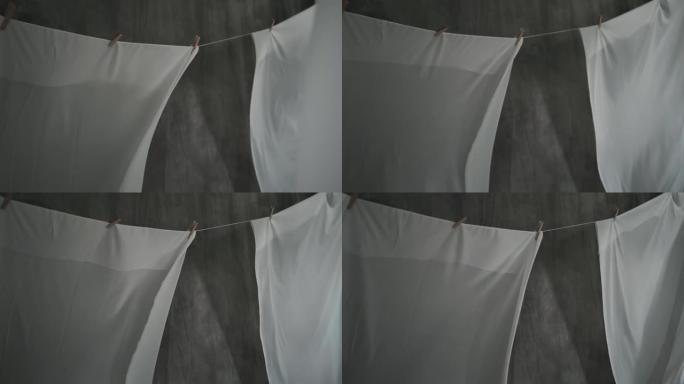 白色织物床单在绳子上的衣夹上干燥。波浪织物在风中飘动。抽象灰色背景。