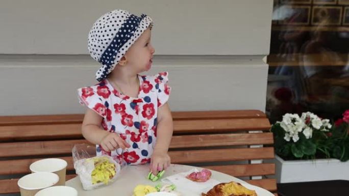 一个穿着彩色连衣裙和圆点帽子的小女孩在城市街头咖啡馆吃彩色蛋糕时弄脏了