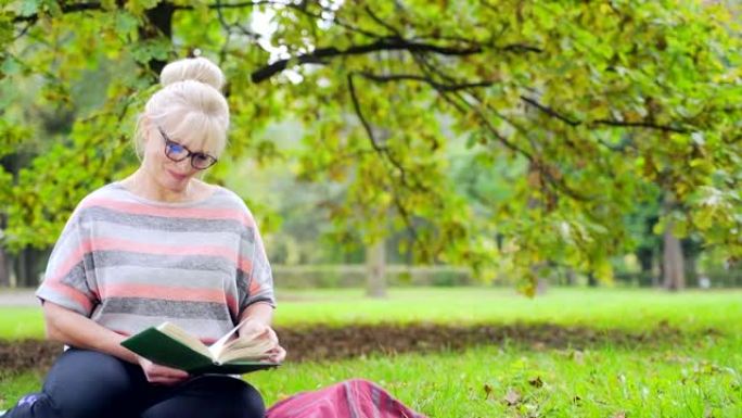 肖像快乐的高级女人坐在公园的草坪上，手里拿着一本书。