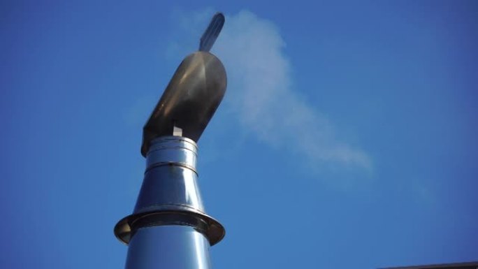 白烟从烟囱上的不锈钢风定向罩上飞来，随风轻轻旋转。带有帽子尖端的炉子的烟道管安装在蓝色天空的金属屋顶