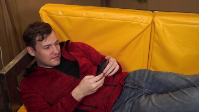 穿着便服的年轻人躺在黄色沙发上。成年男性休息并在手机中浏览各种新闻