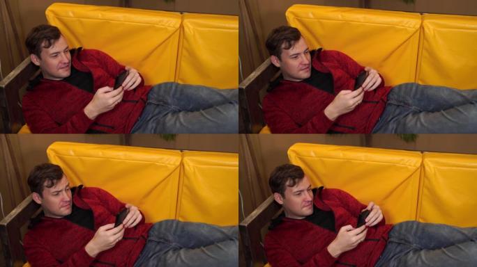 穿着便服的年轻人躺在黄色沙发上。成年男性休息并在手机中浏览各种新闻