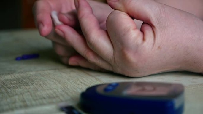 患有糖尿病的老年妇女在家中使用血糖仪测量和检查血糖。女人用棉絮止住手指上的血。一个血糖仪在桌子上。选