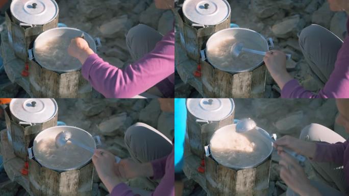 一名妇女在远足时在山中烹饪时，用大锅中的钢包搅拌煮粥。