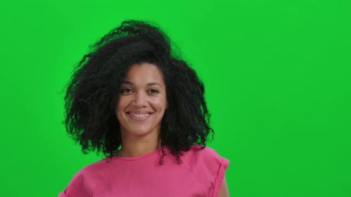 年轻女性非洲裔美国人的肖像看着镜头，微笑可爱。卷发的黑人妇女在工作室的绿色屏幕上摆姿势。特写。慢动作