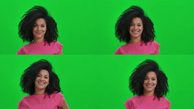 年轻女性非洲裔美国人的肖像看着镜头，微笑可爱。卷发的黑人妇女在工作室的绿色屏幕上摆姿势。特写。慢动作