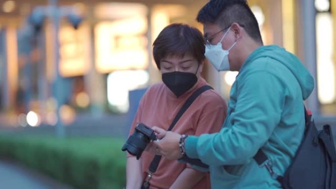 亚洲中国中年夫妇游客晚上在繁忙的吉隆坡城市街道用相机拍照，霓虹灯带面罩