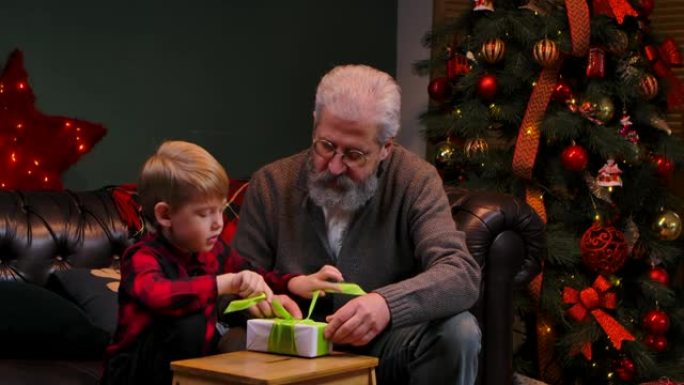 优雅的老人和小男孩打包礼品盒并系蝴蝶结。祖父和孙子坐在装饰房间的沙发上，靠近一棵发光的圣诞树。家庭度