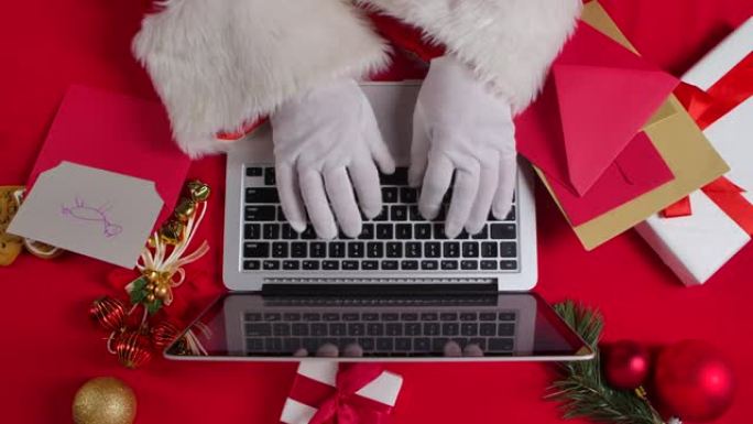 顶视图圣诞老人戴着白手套的手在红色新年装饰桌的键盘笔记本电脑上打字。圣诞老人浏览孩子们的来信，并在互