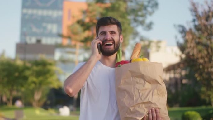 完美的晴天迷人的男人带着装满食物的环保袋走在街上，他在智能手机上讲话，享受时光