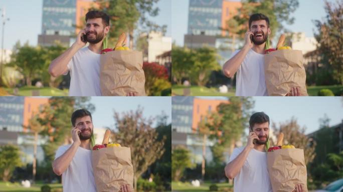 完美的晴天迷人的男人带着装满食物的环保袋走在街上，他在智能手机上讲话，享受时光