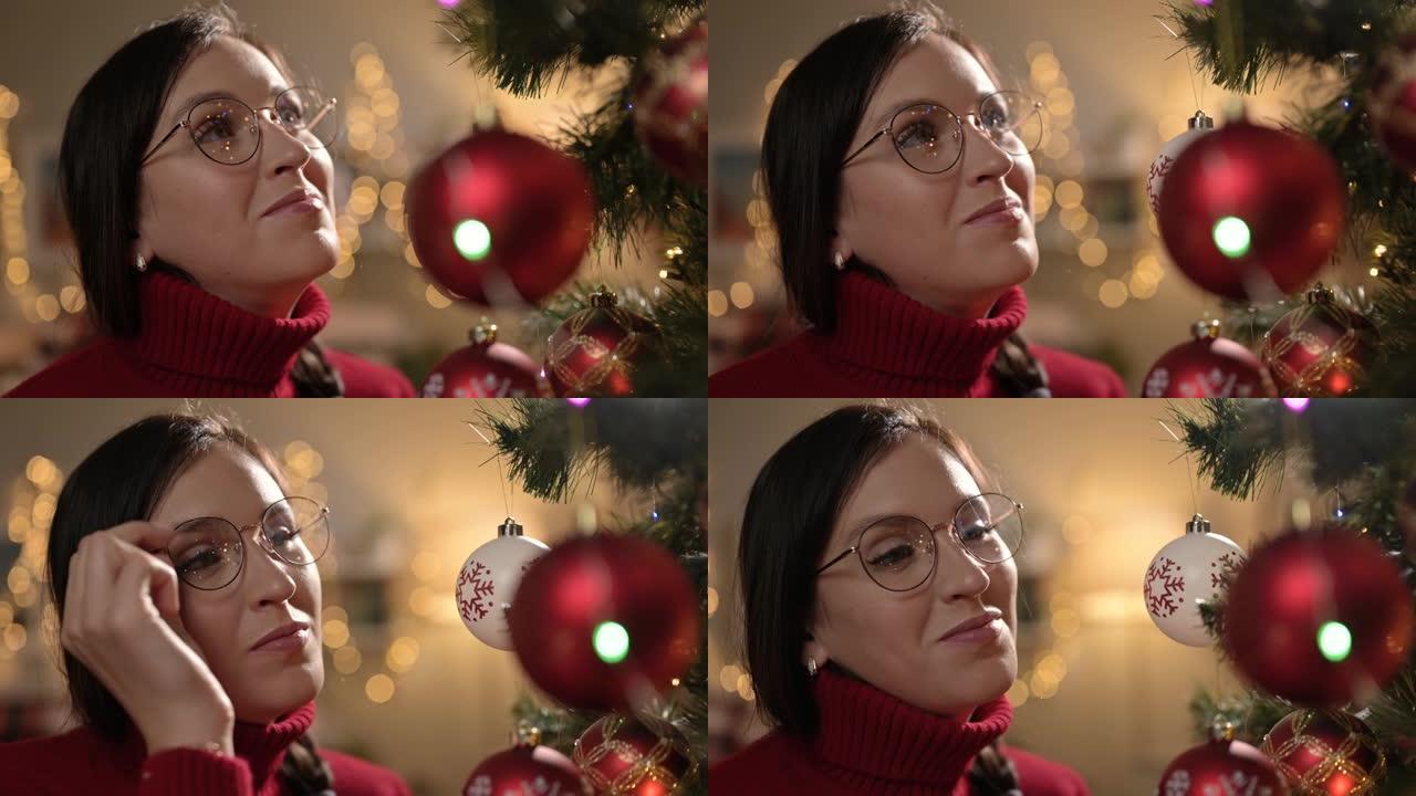 平安夜。快乐的女人看着圣诞树并欢欣鼓舞，花环的圣诞灯映在眼镜上。特写