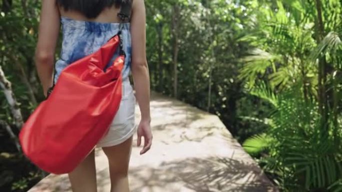 活跃的黑发亚洲女人，带着防水袋独自走在木头的人行道上。女性旅行者探索，户外冒险生活方式，湿热，后视图