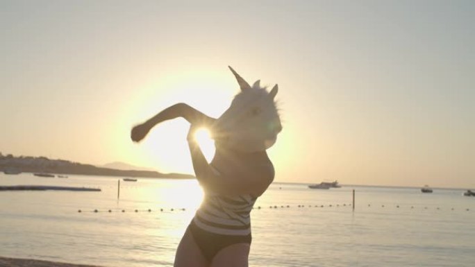 穿着泳衣和独角兽头面具的女人在阳光明媚的海滩上跳舞。戴着独角兽面具的人的有趣舞蹈。快乐的人。