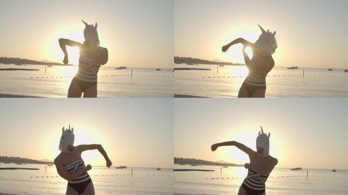 穿着泳衣和独角兽头面具的女人在阳光明媚的海滩上跳舞。戴着独角兽面具的人的有趣舞蹈。快乐的人。