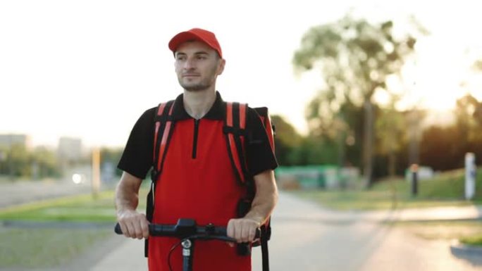 一位戴着眼镜的年轻学生在送货服务中工作，戴着红色背包和棒球帽，在日落时沿着堤岸骑着电动踏板车